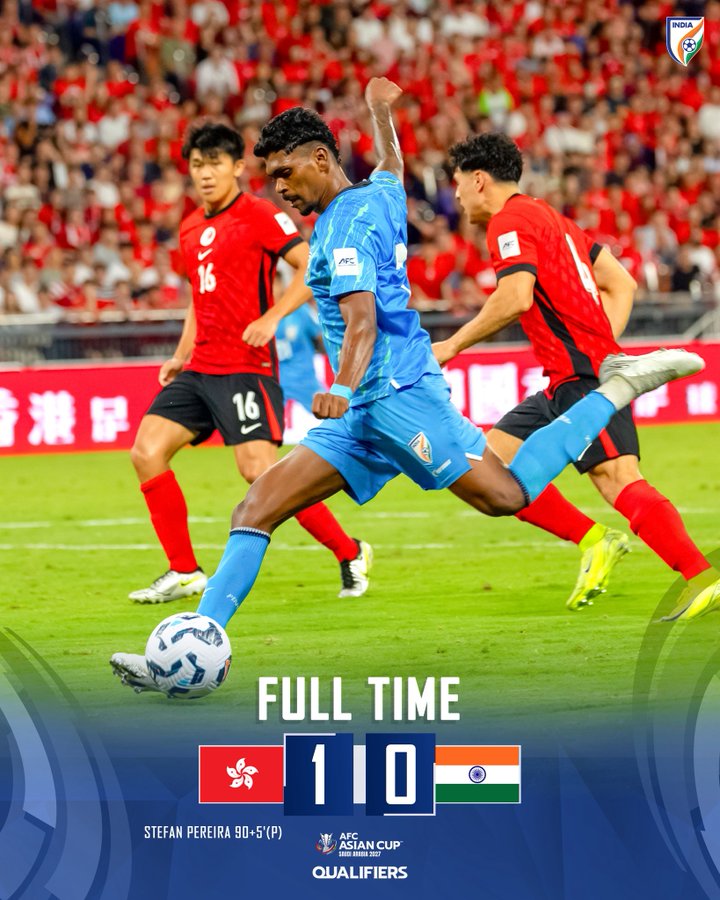 Indian football player in blue kit taking a shot during the AFC Asian Cup 2027 Qualifier against Hong Kong, with the scoreboard showing a 1-0 victory for Hong Kong.
